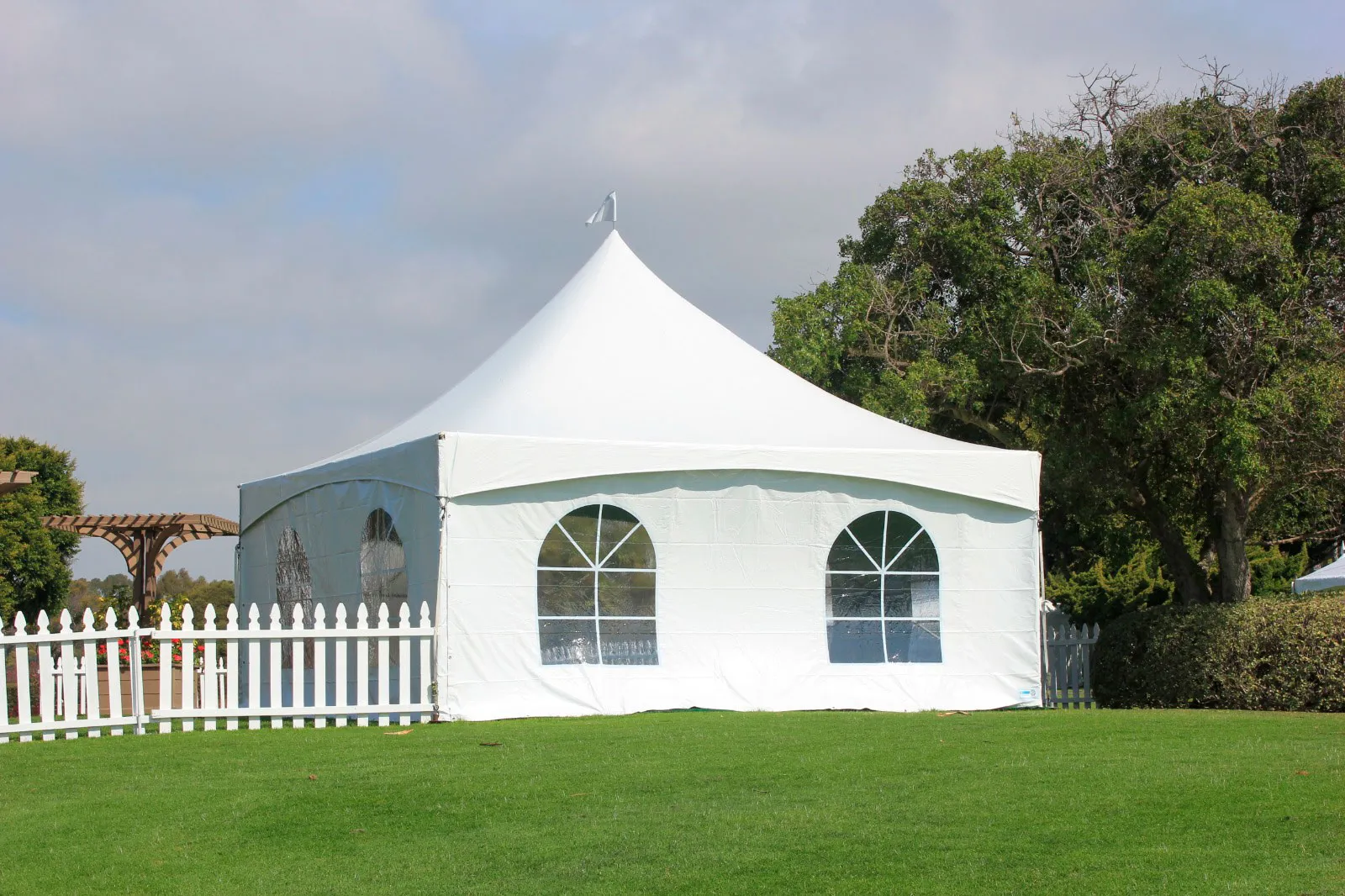 A white gable-ended frame tent with arched window sidewalls behind a white picket fence on a lawn.
