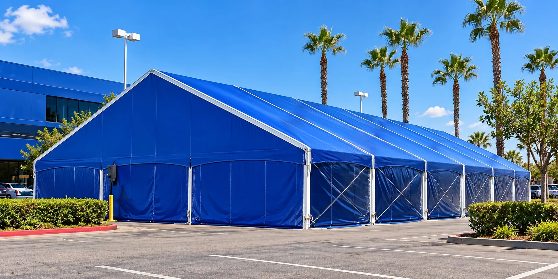 A long blue clearspan structure with a white frame on a paved lot lined with palm trees.
