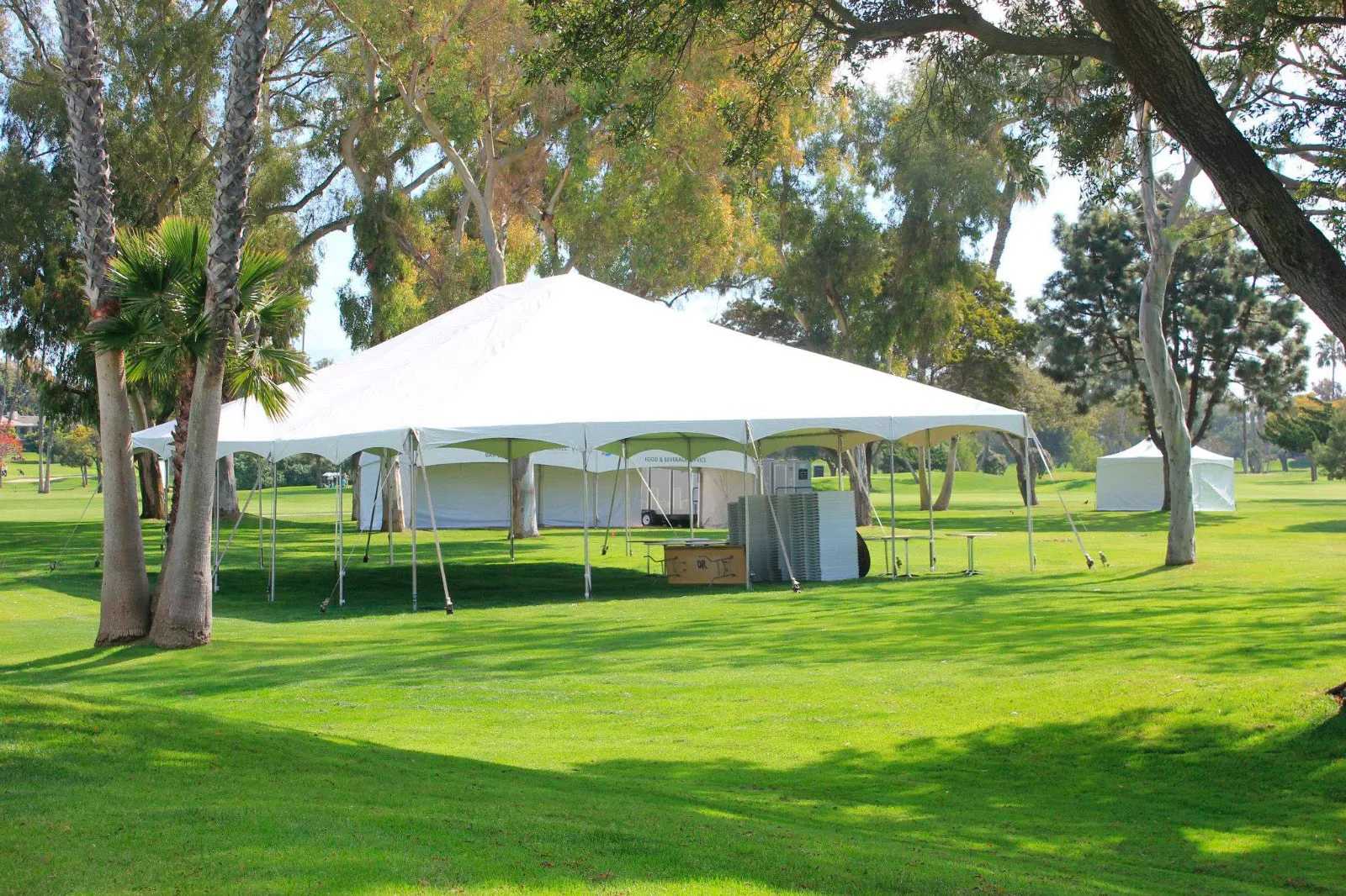 A white frame tent with sidewalls on grass beneath mature trees, with a smaller canopy alongside.