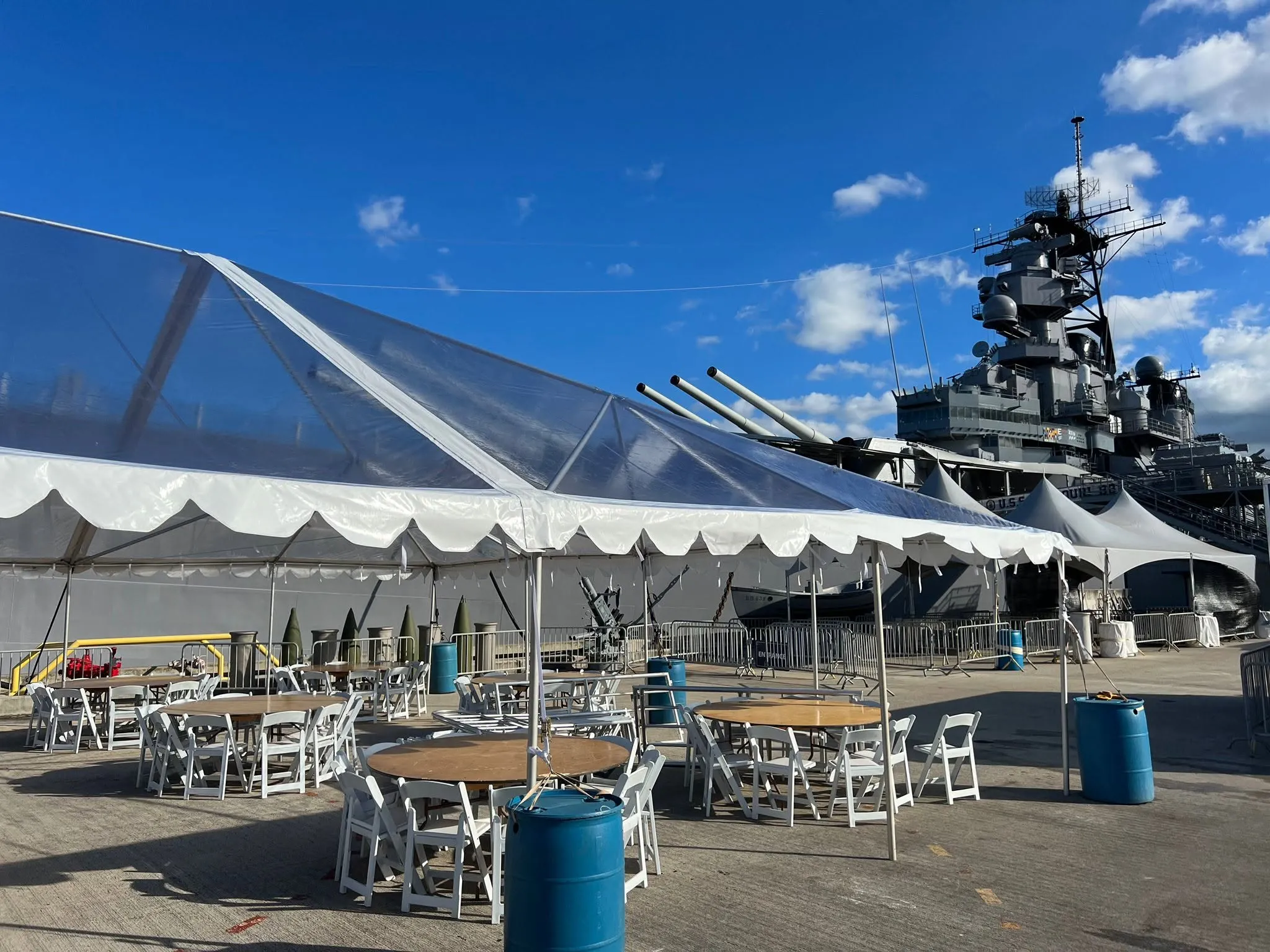 A run of white frame tents with a scalloped valance and a clear-top section, set with tables and chairs beside a moored battleship.
