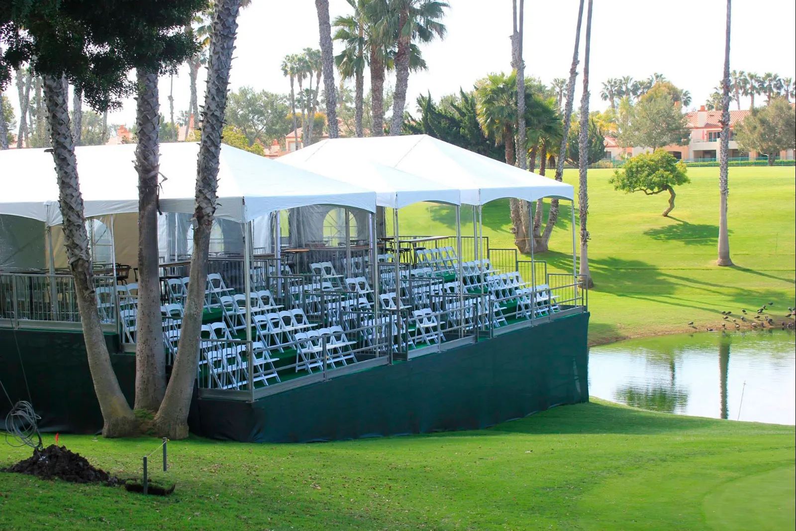 A long white frame tent with clear sidewalls and tiered seating beside a pond and palm trees.