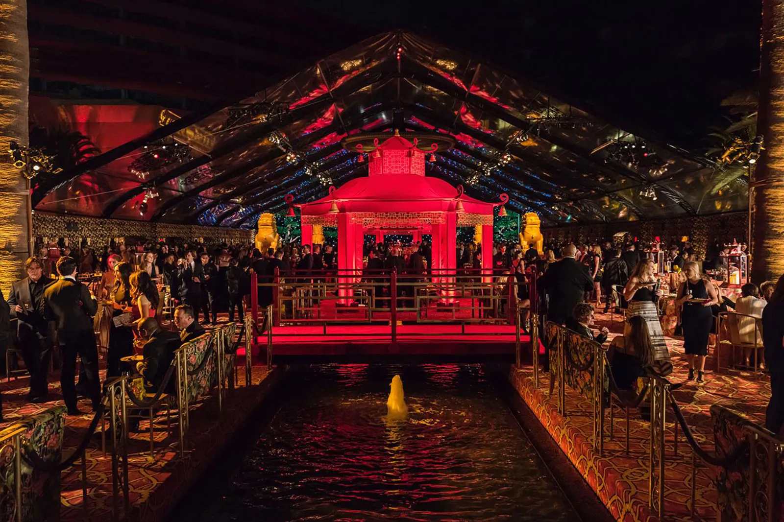 A large pavilion lit deep red at night, its structure reflected in water in the foreground.