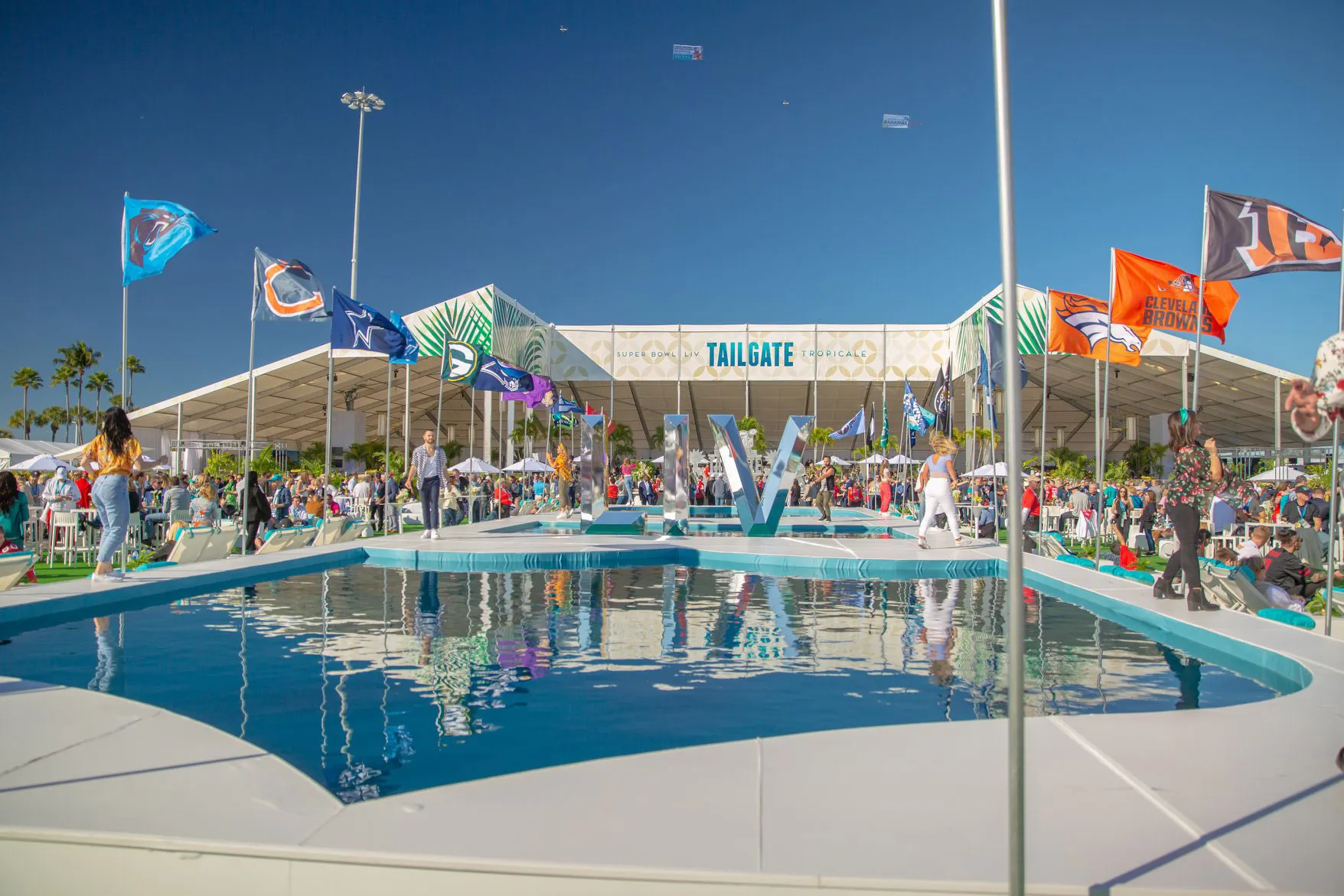 A large white clearspan structure with an open frontage, fronted by a pool and rows of NFL team flags, under a clear sky.